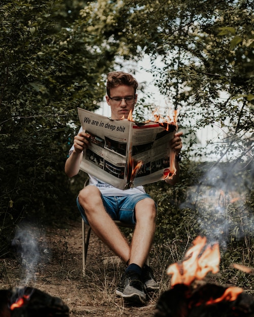 Jovem sentado em uma cadeira segurando e lendo um jornal em chamas