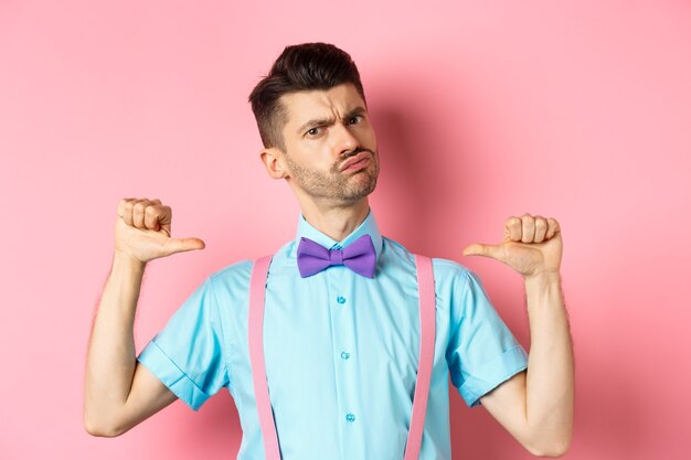 Jovem sério e engraçado tentando parecer legal, apontando para si mesmo para se autopromover, sendo um profissional, em pé sobre um fundo rosa.