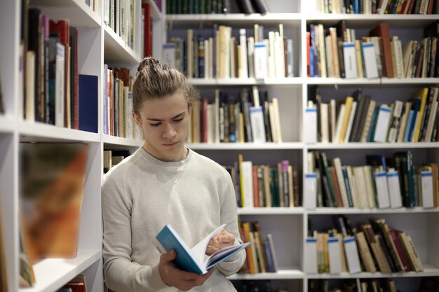 Jovem sério vestindo um suéter em pé na livraria, lendo um trecho de um livro em suas mãos, apoiado em prateleiras brancas cheias de livros