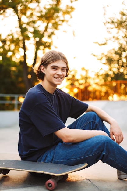 Jovem skatista sorridente em camiseta preta e jeans alegremente olhando na câmera passando tempo com skate no moderno parque de skate
