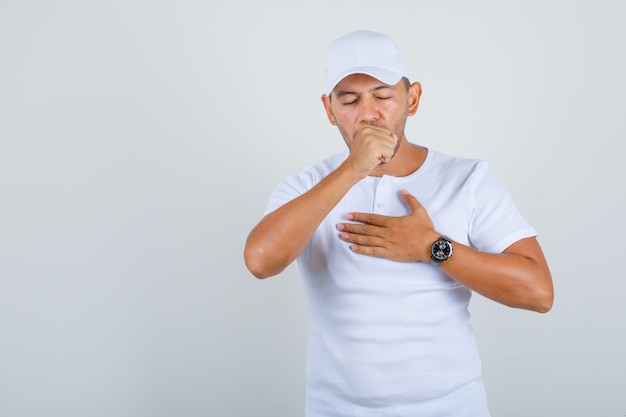 Jovem sofrendo com tosse em t-shirt branca, boné e parecendo doente, vista frontal.