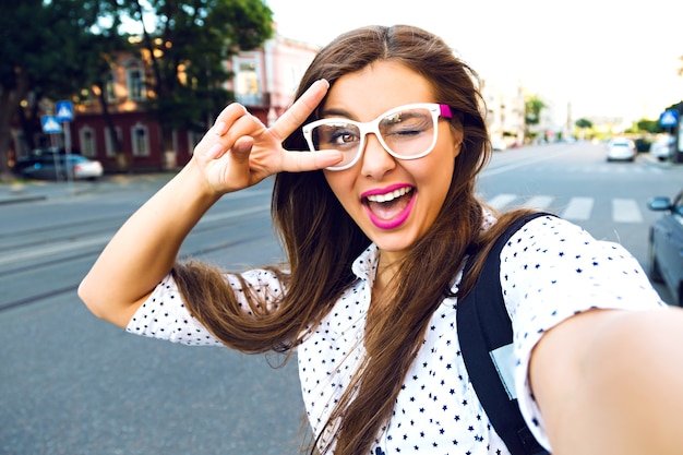 Jovem sorridente adolescente feliz fazendo selfie na rua, cabelos castanhos, maquiagem brilhante e lindos óculos transparentes, viajando sozinha, se divertindo, humor positivo, alegria, férias