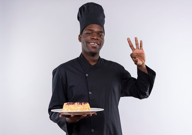 Jovem sorridente afro-americana com uniforme de chef segurando bolo no prato e gesticulando com a mão ok sinal em branco com espaço de cópia
