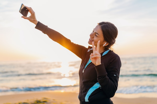 Jovem sorridente atraente magro fazendo exercícios de esporte na praia ao nascer do sol de manhã em roupas esportivas, estilo de vida saudável, ouvindo música em fones de ouvido, fazendo selfie foto no telefone com humor positivo