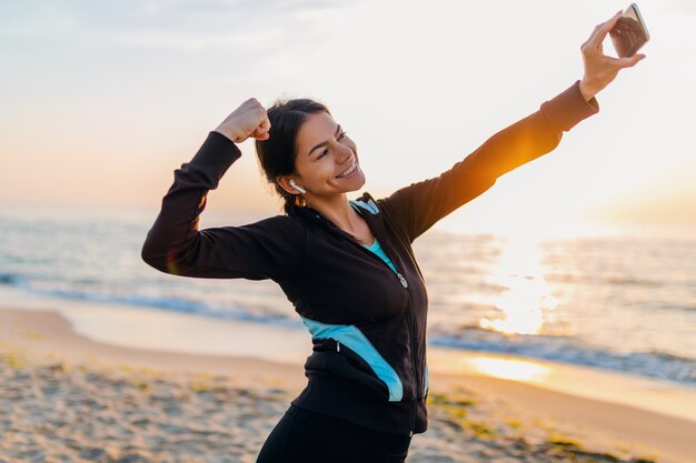 Jovem sorridente atraente magro fazendo exercícios de esporte na praia ao nascer do sol de manhã em roupas esportivas, estilo de vida saudável, ouvindo música em fones de ouvido, fazendo selfie foto no telefone parecendo forte