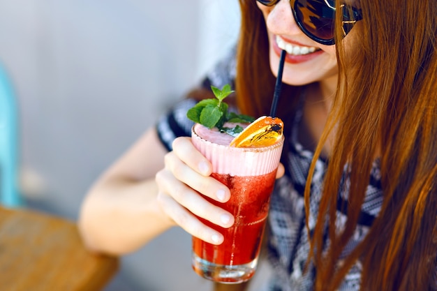 Jovem sorridente bebendo um saboroso coquetel doce, incrível dia relaxante, saborosa limonada, elegante vestido e óculos escuros, terraço ao ar livre.