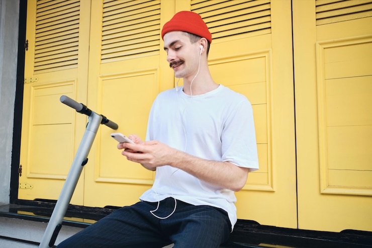 Foto grátis jovem sorridente bigode de camiseta e chapéu vermelho sentado com e-scooter enquanto felizmente usando celular com fundo amarelo na rua da cidade