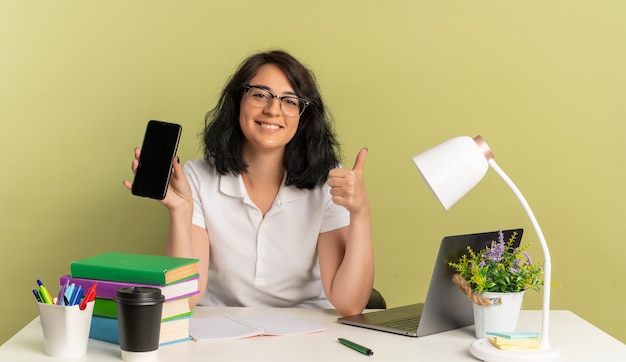 Jovem sorridente, bonita caucasiana colegial de óculos, senta-se na mesa com as ferramentas da escola, polegares para cima e segura o telefone isolado no espaço verde com espaço de cópia