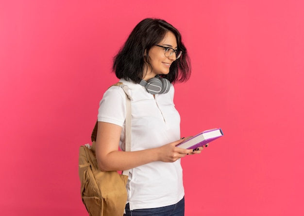 Jovem, sorridente, bonita, caucasiana, estudante, usando óculos, fones de ouvido e bolsa traseira, segurando de lado o livro e o caderno rosa com espaço de cópia