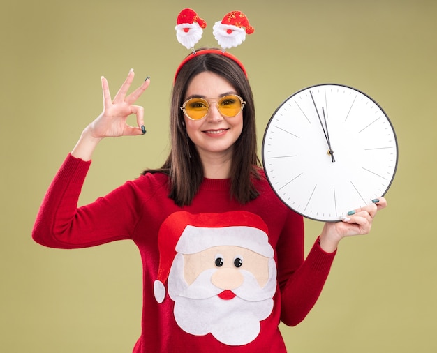 Jovem sorridente, bonita caucasiana, vestindo uma blusa de Papai Noel e bandana com óculos segurando um relógio, olhando para a câmera, fazendo sinal de ok