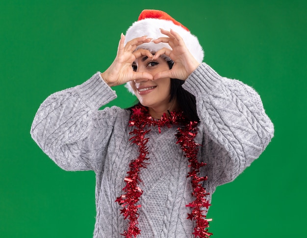 Foto grátis jovem sorridente caucasiana com chapéu de natal e guirlanda de ouropel no pescoço, olhando para a câmera fazendo um sinal de coração na frente do olho isolado no fundo verde