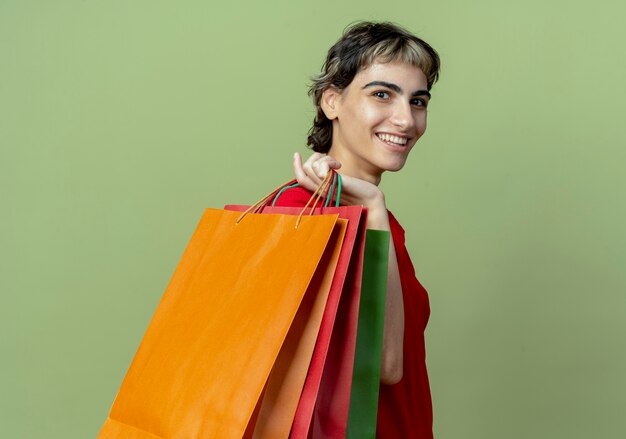 Jovem sorridente caucasiana com corte de cabelo de duende em pé na vista de perfil segurando sacolas de compras no ombro isoladas em fundo verde oliva com espaço de cópia