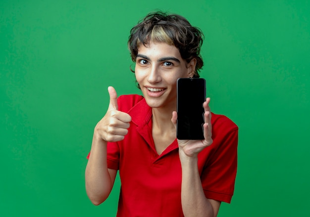 Jovem sorridente caucasiana com corte de cabelo de duende segurando um telefone celular e aparecendo o polegar isolado em um fundo verde com espaço de cópia