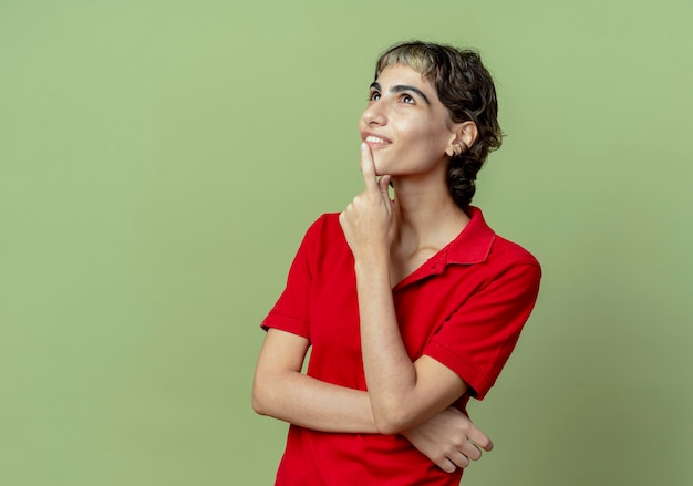 Jovem sorridente caucasiana com corte de cabelo de duende tocando o queixo e olhando para cima isolada em fundo verde oliva com espaço de cópia