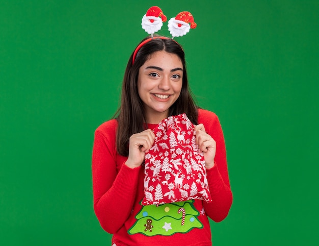 Jovem sorridente caucasiana com fita de Papai Noel segurando uma sacola de presente de Natal isolada em um fundo verde com espaço de cópia