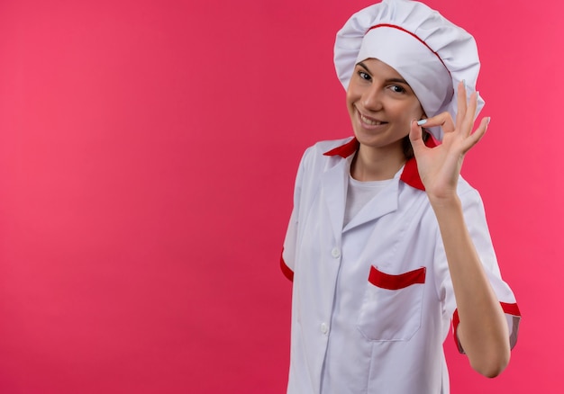 Jovem sorridente caucasiana cozinheira com uniforme de chef gesticula com a mão ok e olha para a câmera rosa com espaço de cópia