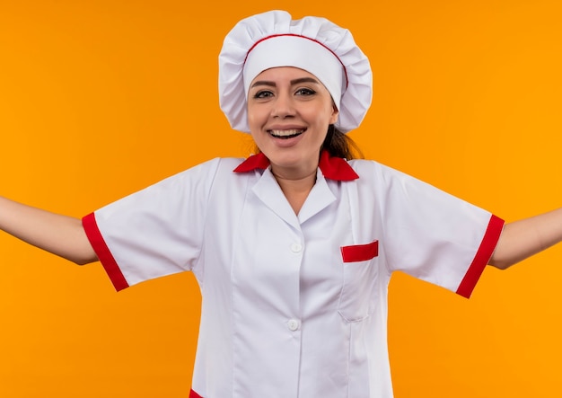 Jovem sorridente caucasiana cozinheira com uniforme de chef olha para a câmera com as mãos abertas, isolada na parede laranja com espaço de cópia