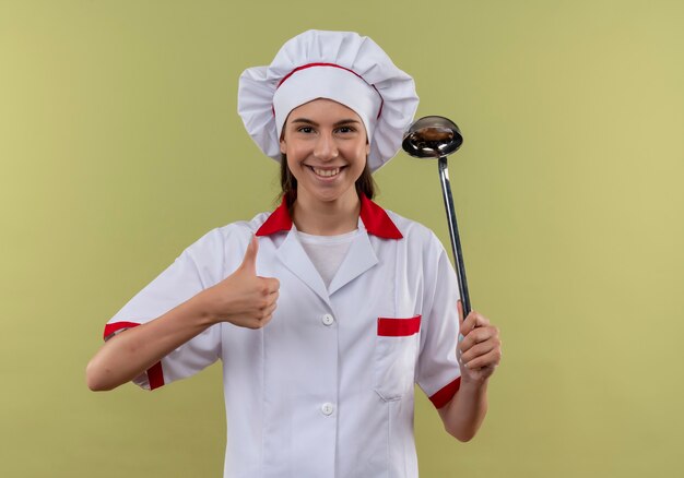 Jovem sorridente caucasiana cozinheira com uniforme de chef segura a concha e o polegar para cima, isolado em um fundo verde com espaço de cópia