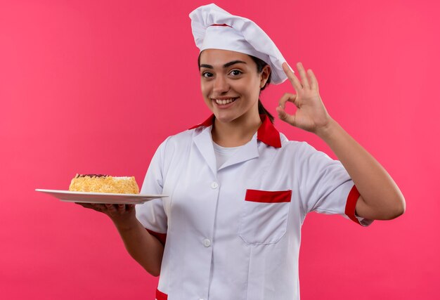 Jovem sorridente caucasiana cozinheira com uniforme de chef segura bolo no prato e gesticula sinal de mão ok isolado na parede rosa com espaço de cópia