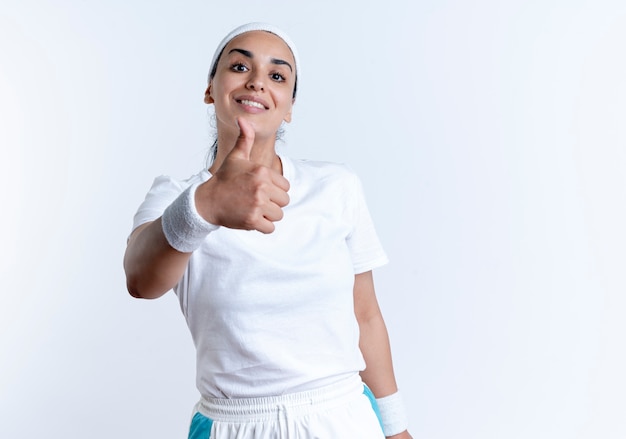 Jovem, sorridente, caucasiana, esportiva, usando fita para a cabeça e pulseiras com os polegares para cima, isolado no espaço em branco com espaço de cópia