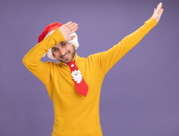 Jovem sorridente, caucasiano, usando um chapéu de Natal e gravata, olhando para a câmera, fazendo um gesto levemente isolado no fundo roxo