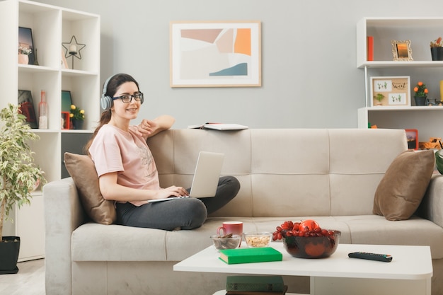 Foto grátis jovem sorridente com laptop usando fones de ouvido e óculos, sentada no sofá atrás da mesa de centro na sala de estar