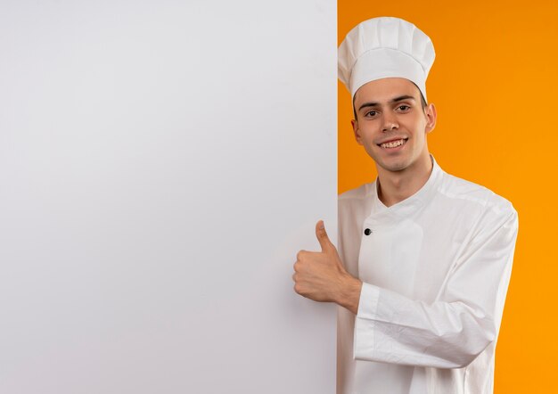 Jovem sorridente com uniforme de chef e segurando uma parede branca com o polegar para cima