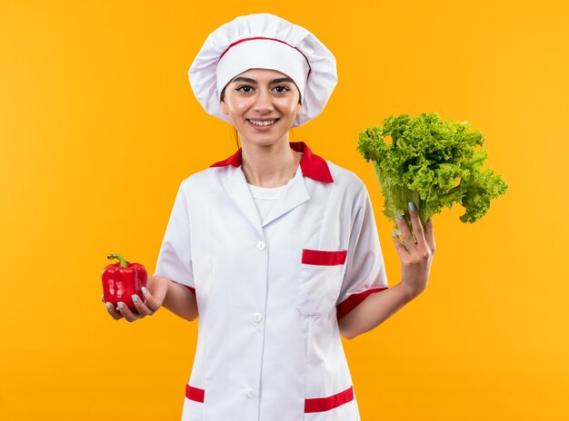 Jovem sorridente com uniforme de chef segurando pimenta com salada isolada na parede laranja