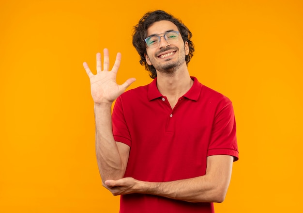 Jovem sorridente de camisa vermelha e óculos ópticos levanta a mão e parece isolado na parede laranja