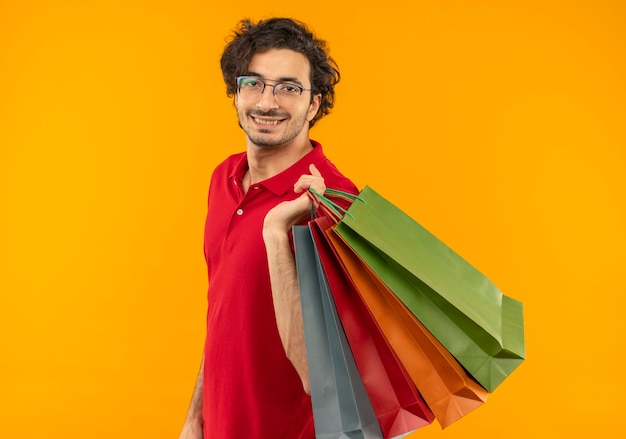 Jovem sorridente, de camisa vermelha e óculos ópticos, segura sacos de papel multicolorido e parece isolado em uma parede laranja