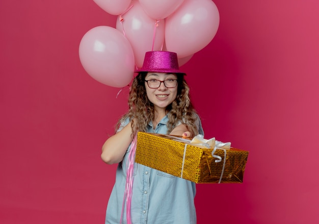 Jovem sorridente de óculos e chapéu rosa segurando nalloons e segurando uma caixa de presente para a câmera isolada no rosa