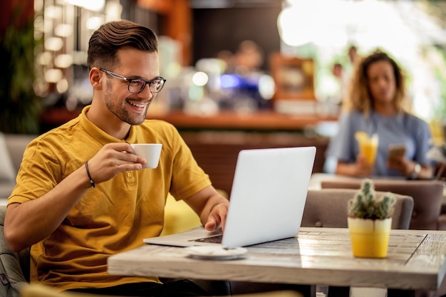 Jovem sorridente digitando em um computador enquanto navega na internet e bebe café em um bar