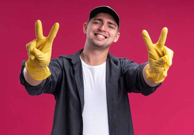 Jovem sorridente e barulhento cara de limpeza vestindo camiseta e boné com luvas, mostrando um gesto de paz isolado na parede rosa