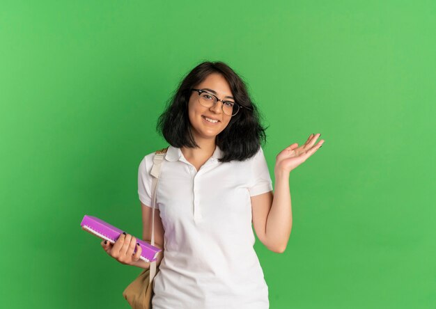 Jovem sorridente e bonita caucasiana colegial de óculos e bolsa nas costas aponta ao lado com a mão segurando livros em verde com espaço de cópia