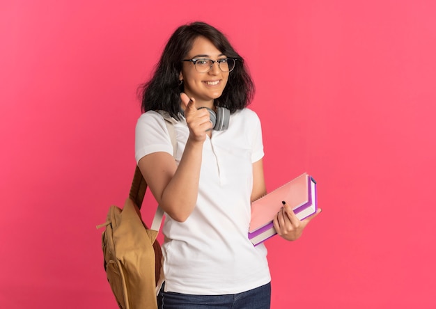 Jovem sorridente e bonita caucasiana colegial de óculos e bolsa traseira com fones de ouvido no pescoço aponta para a câmera segurando livros rosa com espaço de cópia