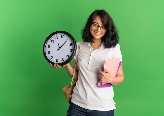 Foto grátis jovem sorridente e bonita caucasiana colegial de óculos e bolsa traseira segura relógio e caderno em verde com espaço de cópia