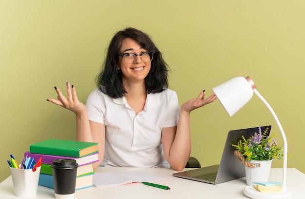 Jovem sorridente e bonita caucasiana colegial de óculos sentada com as mãos levantadas na mesa com ferramentas escolares isoladas em um espaço verde com espaço de cópia