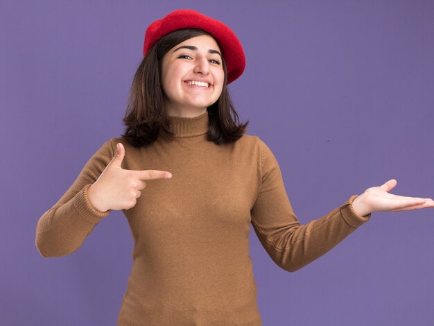 Jovem sorridente e bonita caucasiana com chapéu boina apontando para a mão vazia no roxo