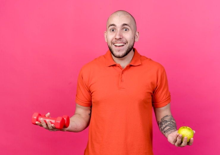 Foto grátis jovem sorridente e desportivo segurando halteres com uma maçã isolada na parede rosa
