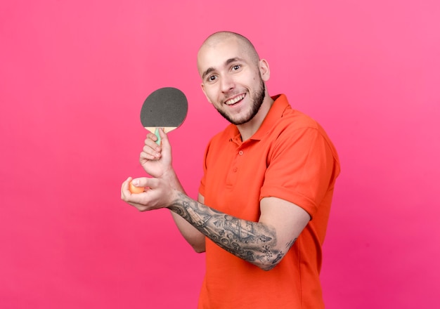 Jovem sorridente e desportivo segurando uma raquete de pingue-pongue com uma bola isolada na parede rosa