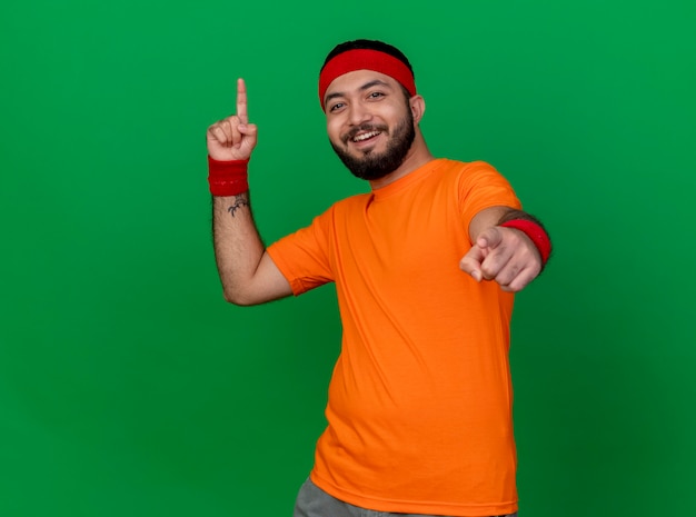 Foto grátis jovem sorridente e desportivo usando bandana e pulseira apontando para cima e para a câmera isolada no verde