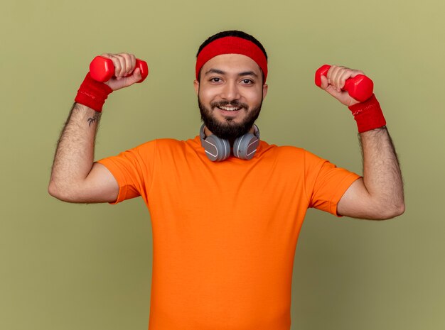 Jovem sorridente e desportivo usando bandana e pulseira com fones de ouvido no pescoço, fazendo exercícios com halteres isolados em fundo verde oliva