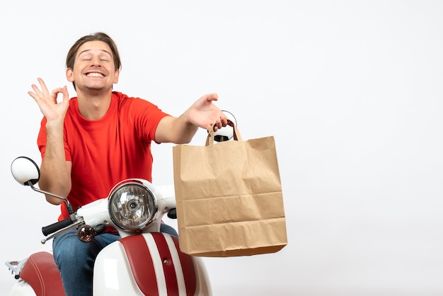 Jovem sorridente e emocional mensageiro de uniforme vermelho sentado na scooter dando uma sacola de papel fazendo um gesto perfeito na parede branca