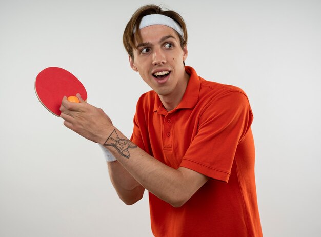 Jovem sorridente e esportivo usando bandana e pulseira segurando uma raquete de pingue-pongue com bola