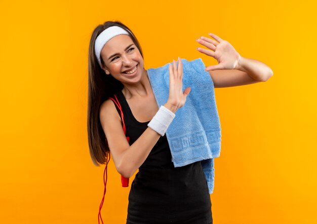 Jovem sorridente e muito esportiva usando bandana e pulseira com uma toalha e pulando corda nos ombros levantando as mãos no espaço laranja