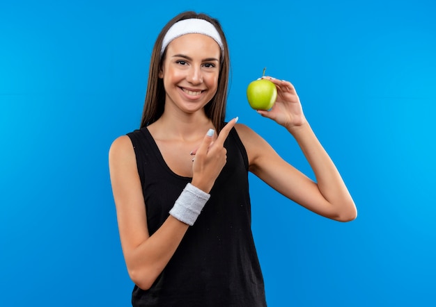 Jovem sorridente e muito esportiva usando bandana e pulseira segurando e apontando para uma maçã isolada no espaço azul
