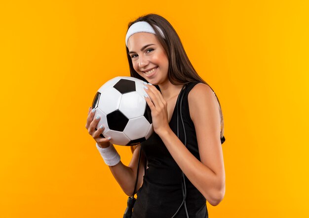 Jovem sorridente e muito esportiva usando bandana e pulseira, segurando uma bola de futebol e pular corda no pescoço, isolada no espaço laranja