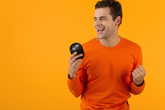 Jovem sorridente elegante com suéter laranja segurando um alto-falante sem fio feliz ouvindo música se divertindo