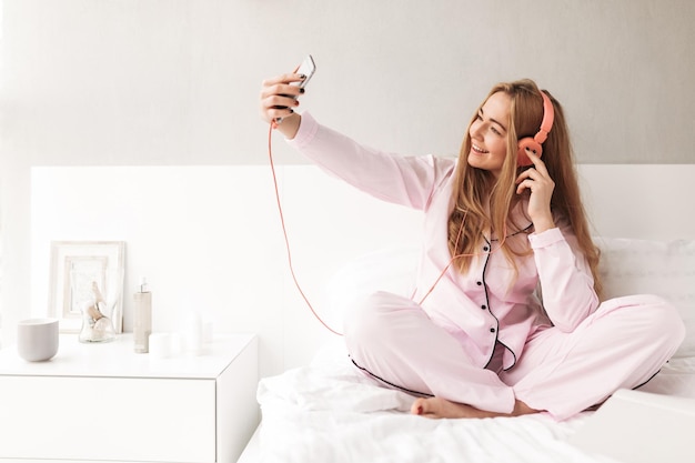 Jovem sorridente em roupas de dormir sentada na cama e ouvindo música em fones de ouvido enquanto tira fotos no celular em casa