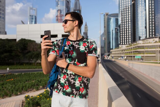 Jovem sorridente em t-shirt e óculos de sol com mochila sonhadoramente olhando de lado enquanto segura o celular na mão com belos arranha-céus da cidade no fundo